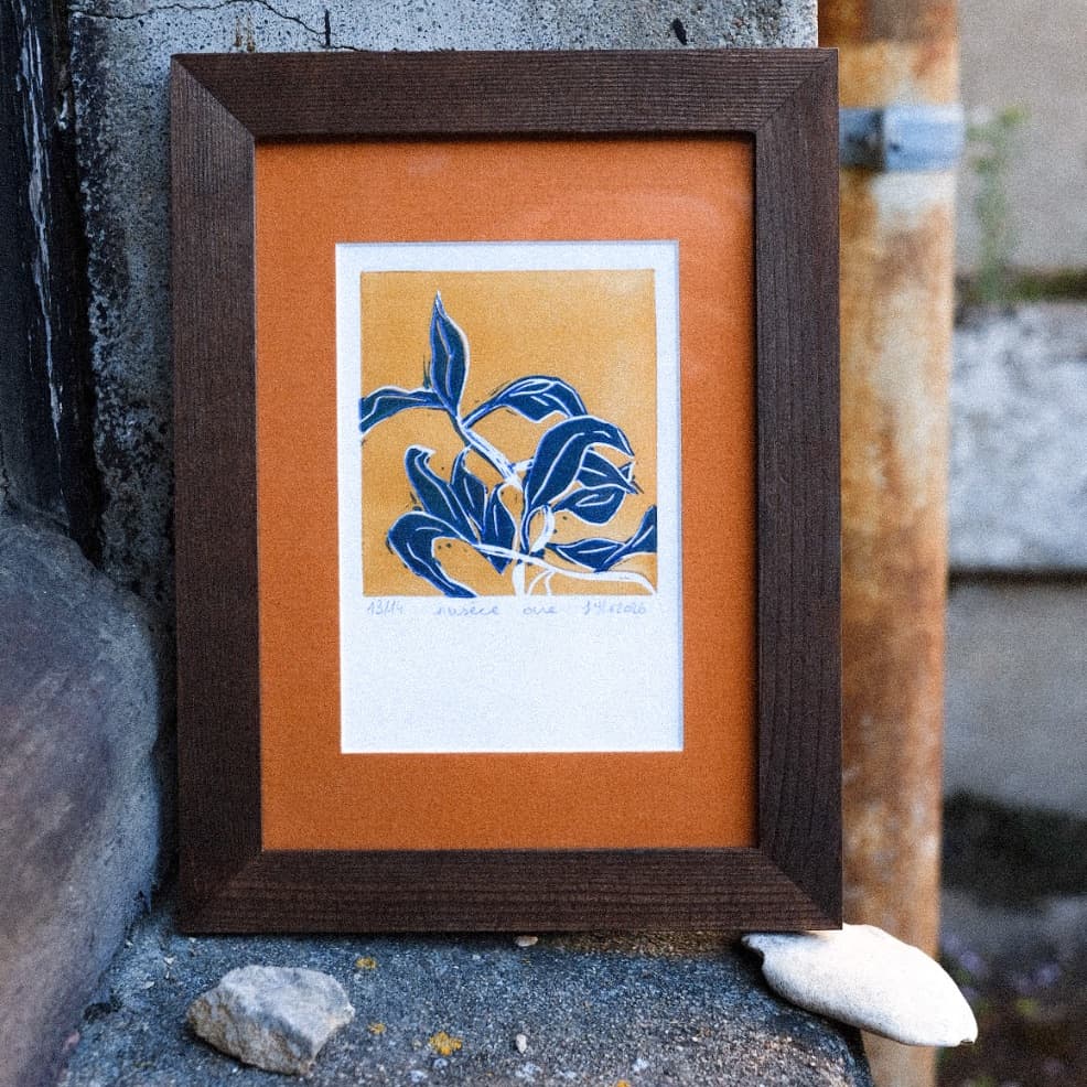 Linogravure Les Misères aux feuilles bleues sur fond ocre, présentée dans un passe-partout terracotta et cadre sombre