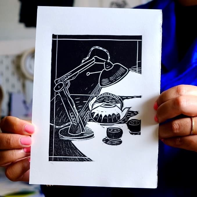 Linogravure tenue en main montrant une lampe de bureau éclairant une théière et deux tasses en noir et blanc.