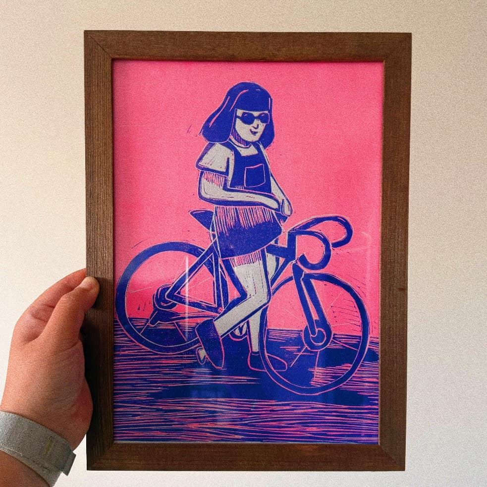 Cadre en bois tenu à la main avec estampe d'une cycliste bleue sur fond rose vif.
