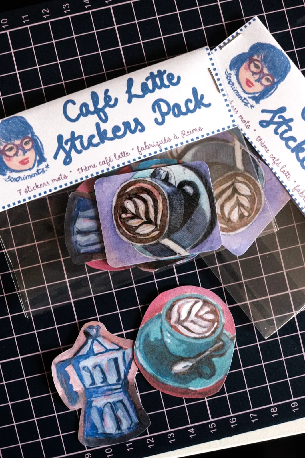 Autocollants illustrés de tasses de café latte et d'une cafetière italienne bleue disposés sur un tapis quadrillé.