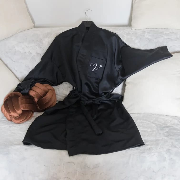Peignoir en satin noir posé à plat avec ceinture nouée et lettre brodée sur la poche.