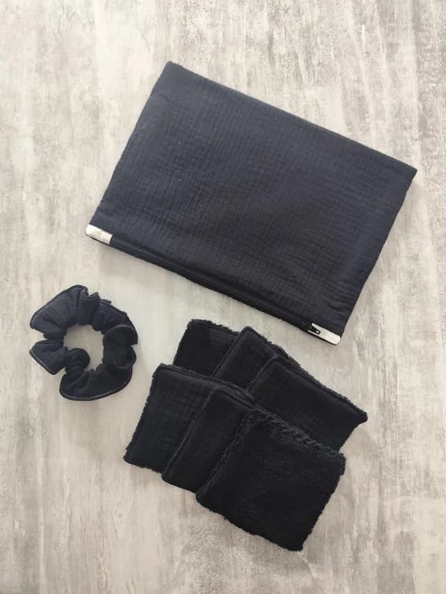Pochette zippée noire, chouchou assorti et lingettes lavables carrées posés à plat sur fond gris.
