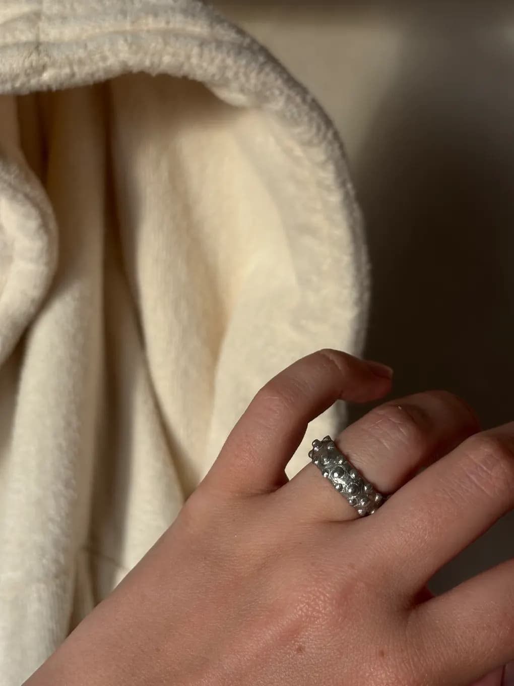 Bague en étain texturée de petites sphères en relief portée à la main devant un tissu écru.