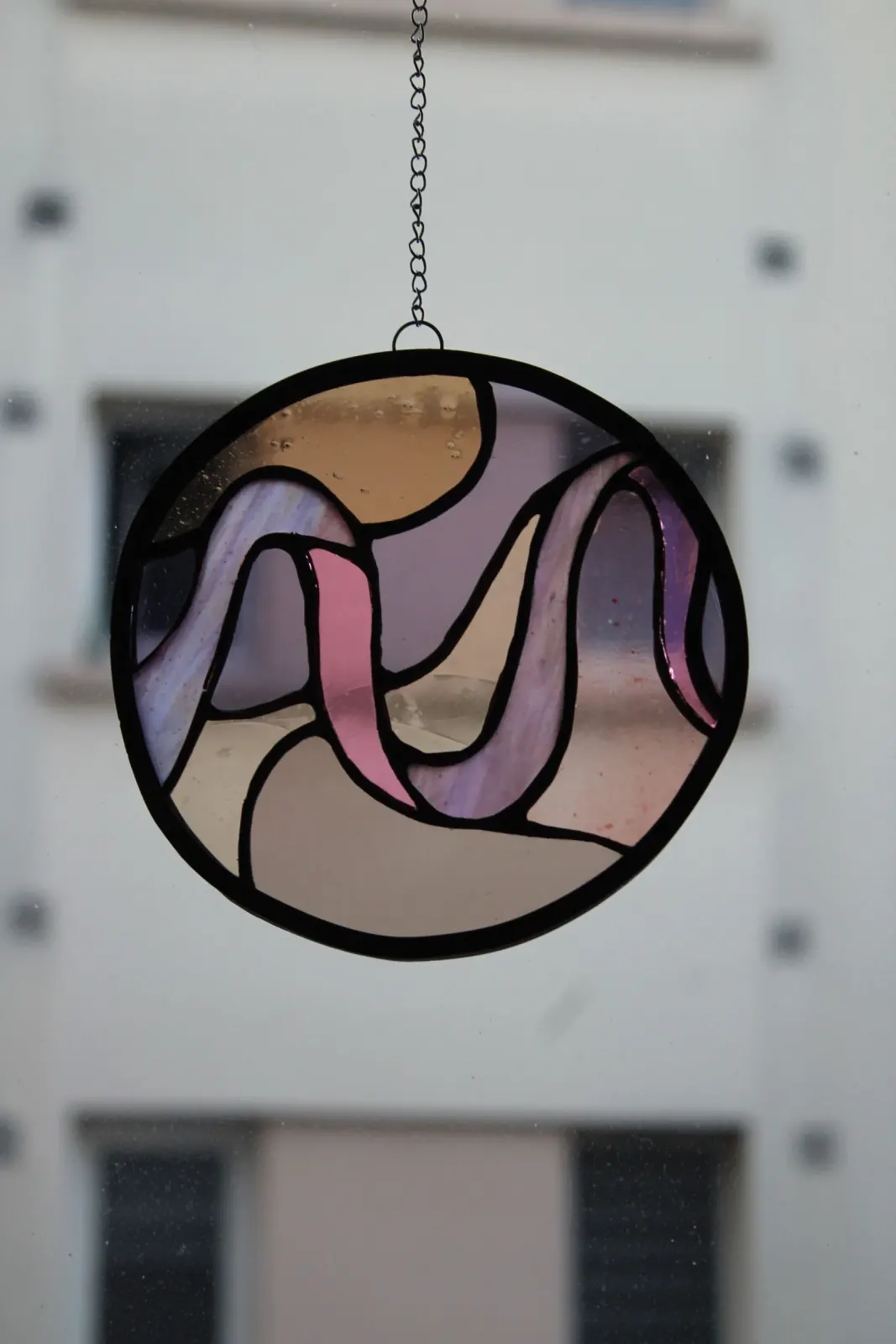 Suspension vitrail circulaire aux formes ondulées rose, violette et dorée devant une fenêtre