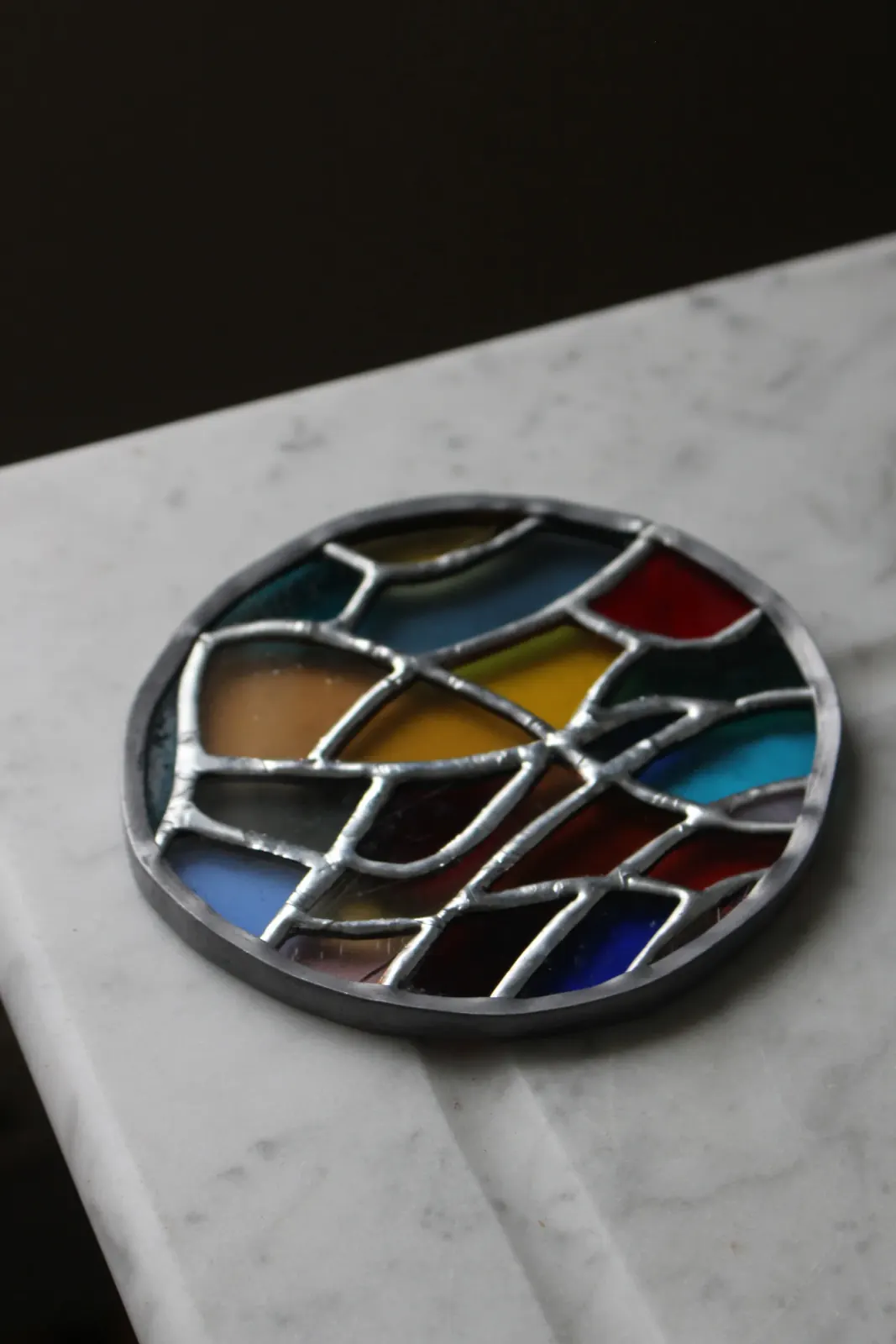 Vitrail rond artisanal posé sur une surface en marbre, vue oblique sur les soudures métalliques.