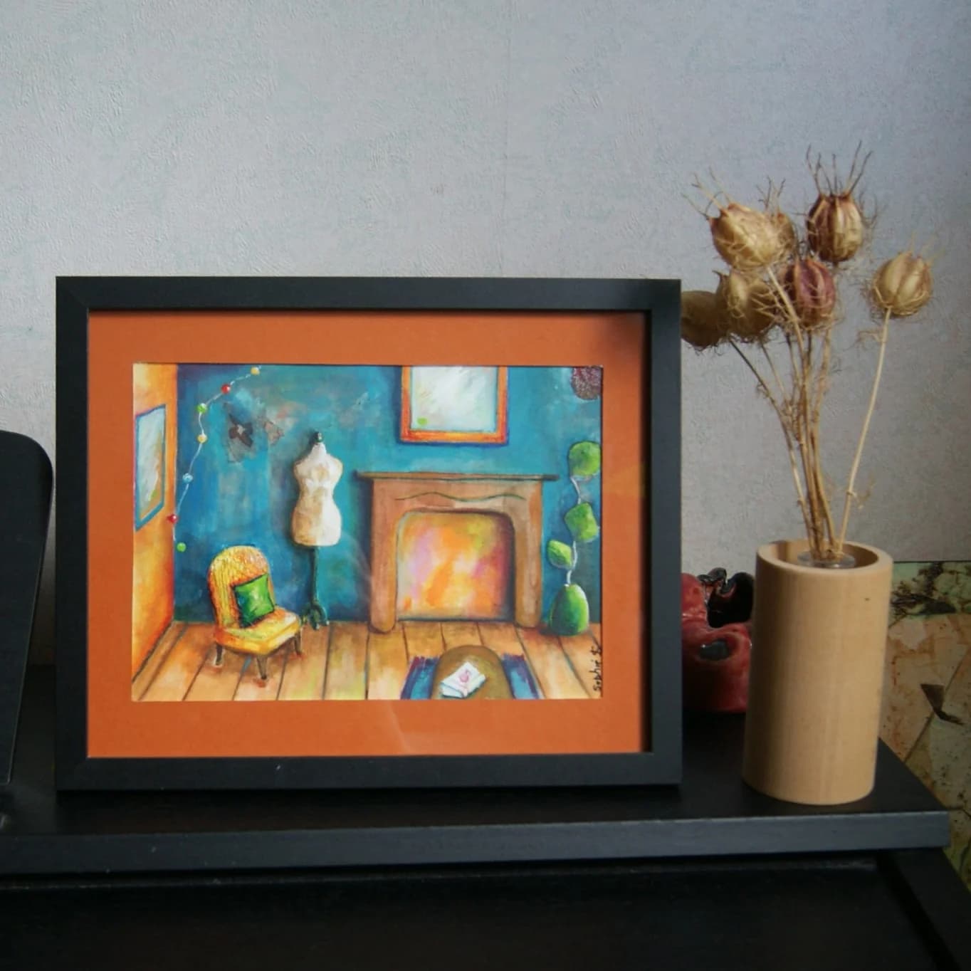 Cadre noir avec passe-partout orange contenant la peinture d'un salon intimiste, posé près d'un vase.