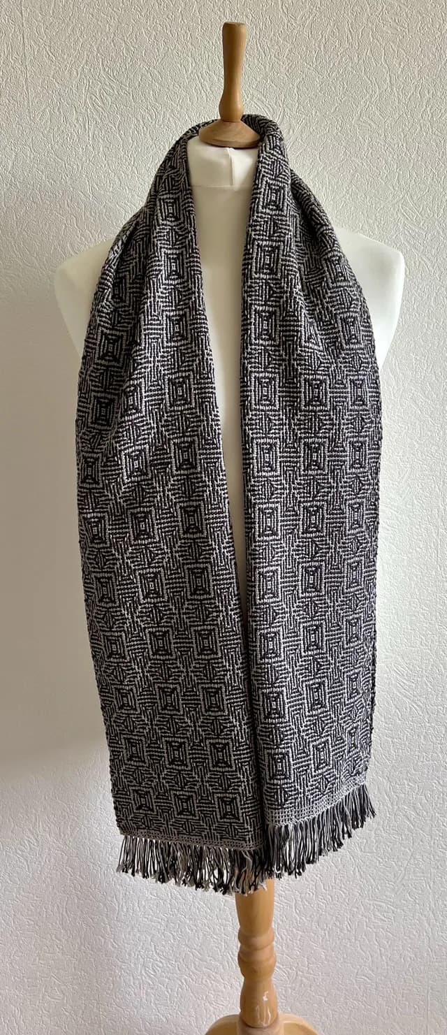 Écharpe tissée noire et grise à motifs géométriques et franges, portée sur un mannequin de couture.