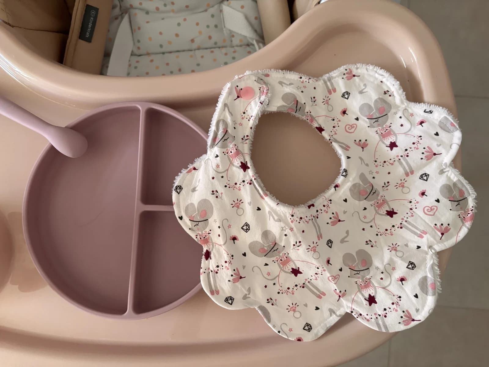 Bavoir en forme de fleur à motifs de souris danseuses posé sur une tablette à côté d'une assiette rose.