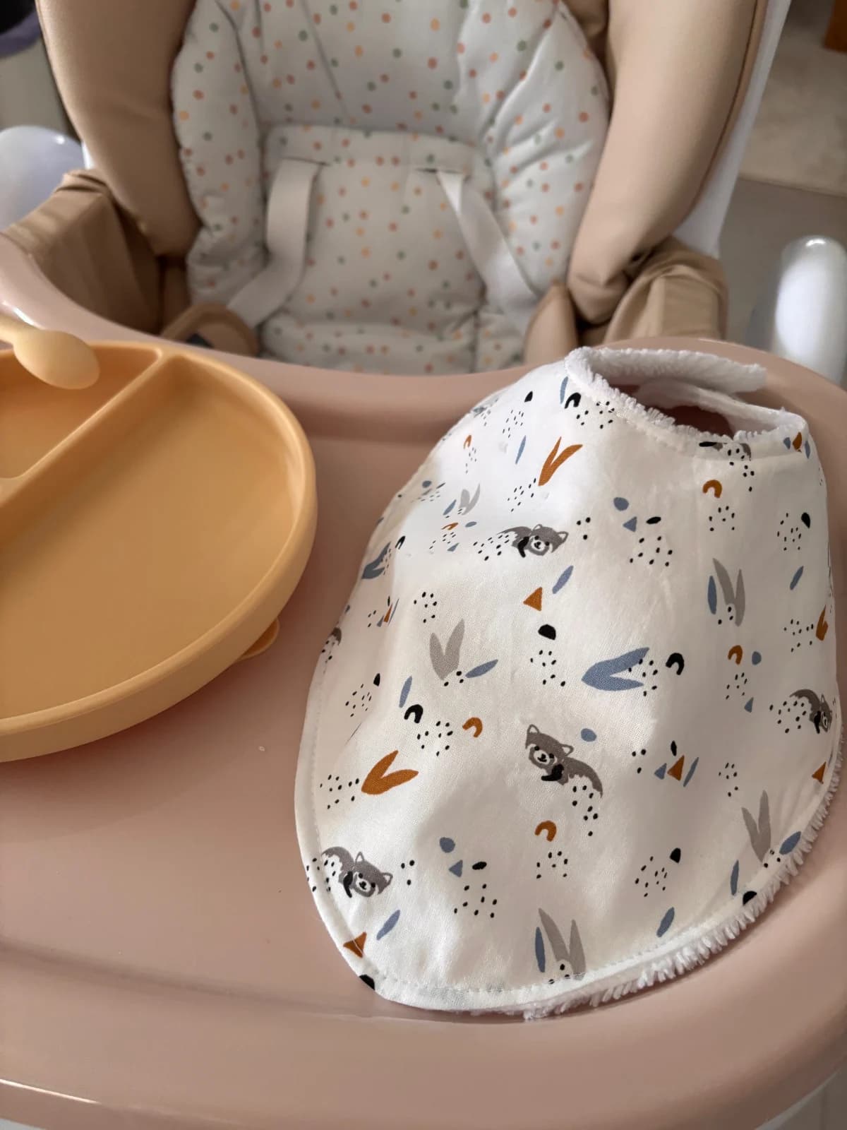Bavoir bandana blanc à motifs d'animaux de la forêt et feuillages posé sur une tablette de chaise haute