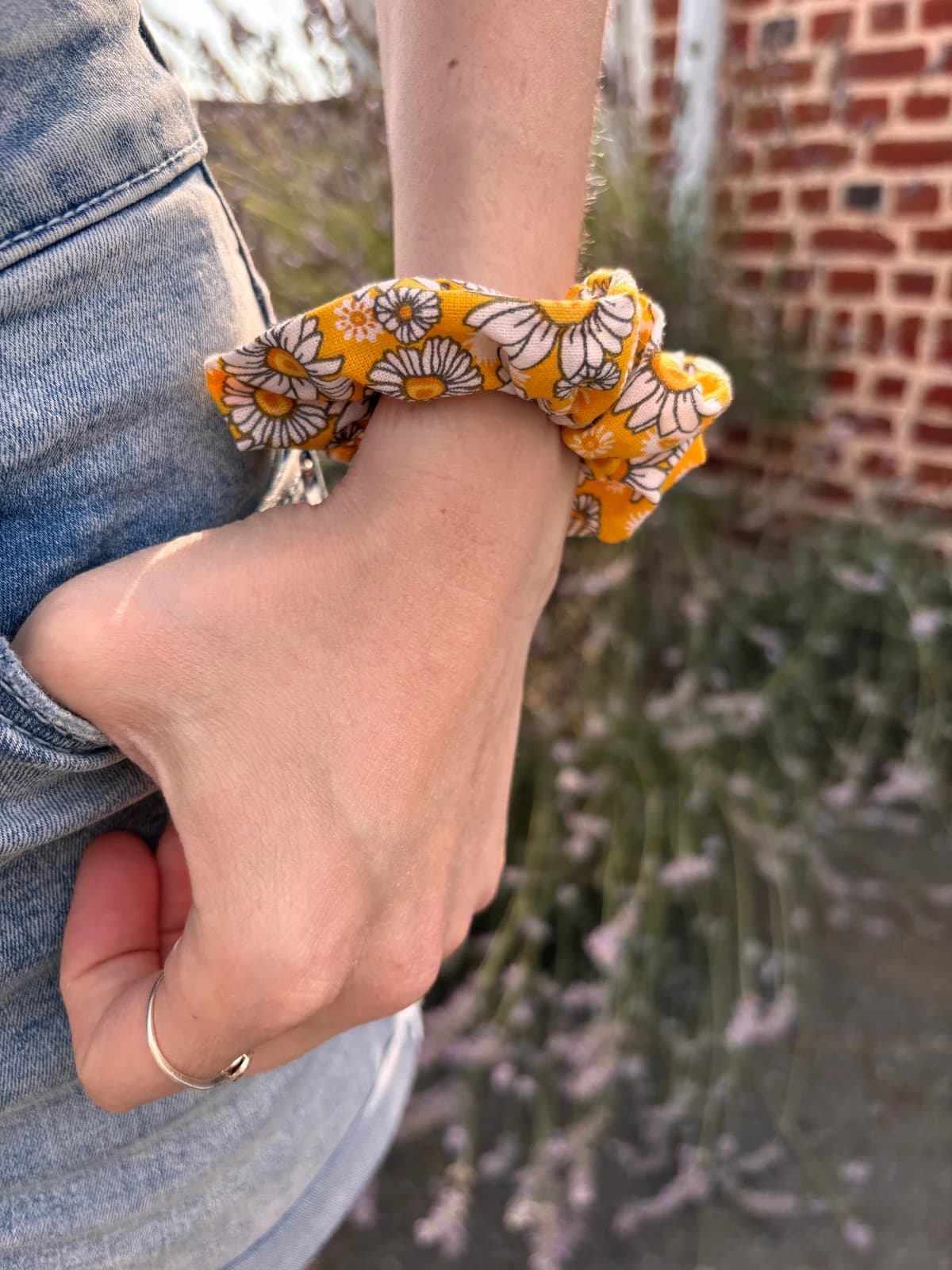 Chouchou en tissu jaune à motifs de marguerites blanches porté au poignet sur un jean.