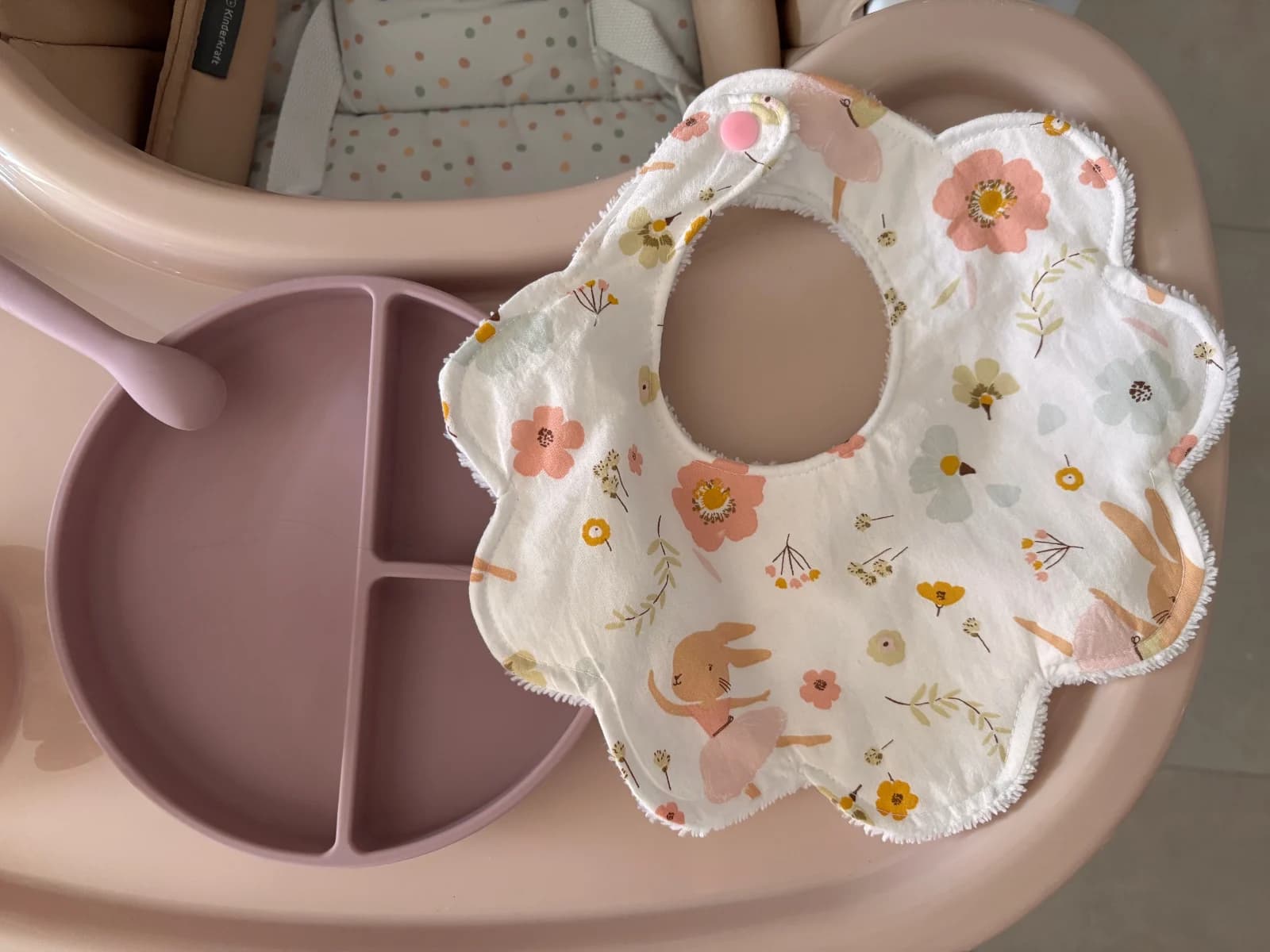 Bavoir en forme de fleur blanc à motifs floraux et lapins, posé à côté d'une assiette sur une tablette rose.