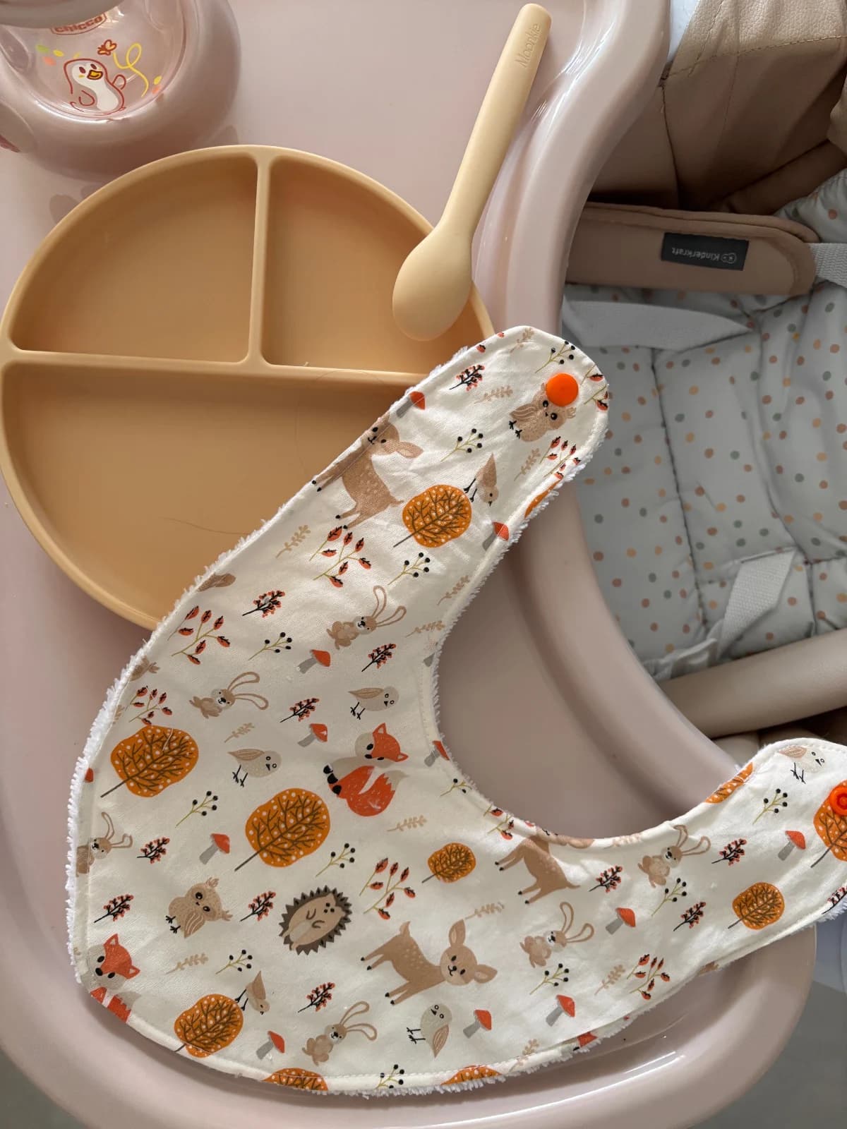 Vue du dessus du bavoir bandana déplié à côté d'une assiette et d'une cuillère beige pour bébé.