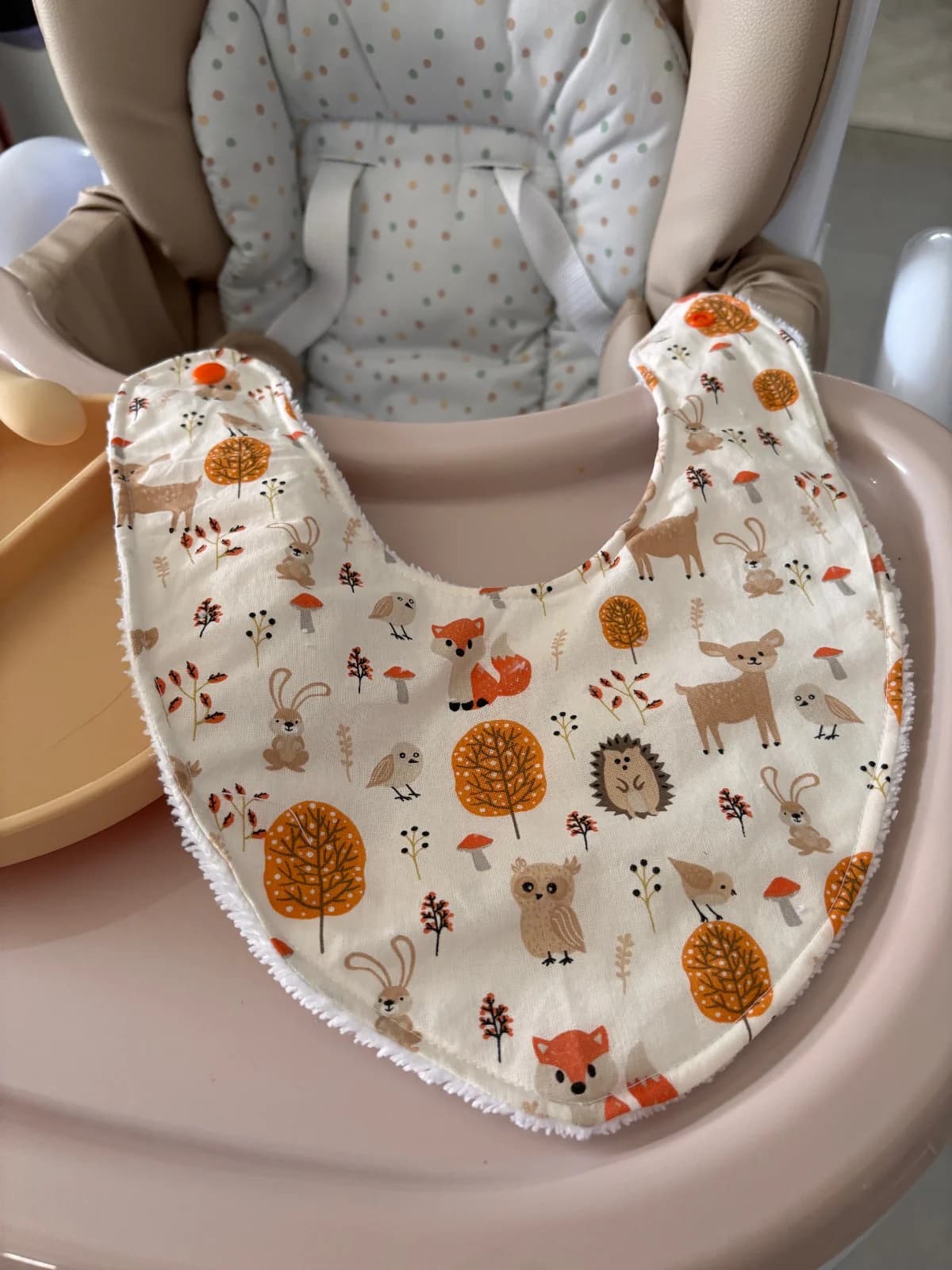 Bavoir bandana ouvert à motifs d'animaux de la forêt dans les tons orange et beige, posé sur une chaise haute.