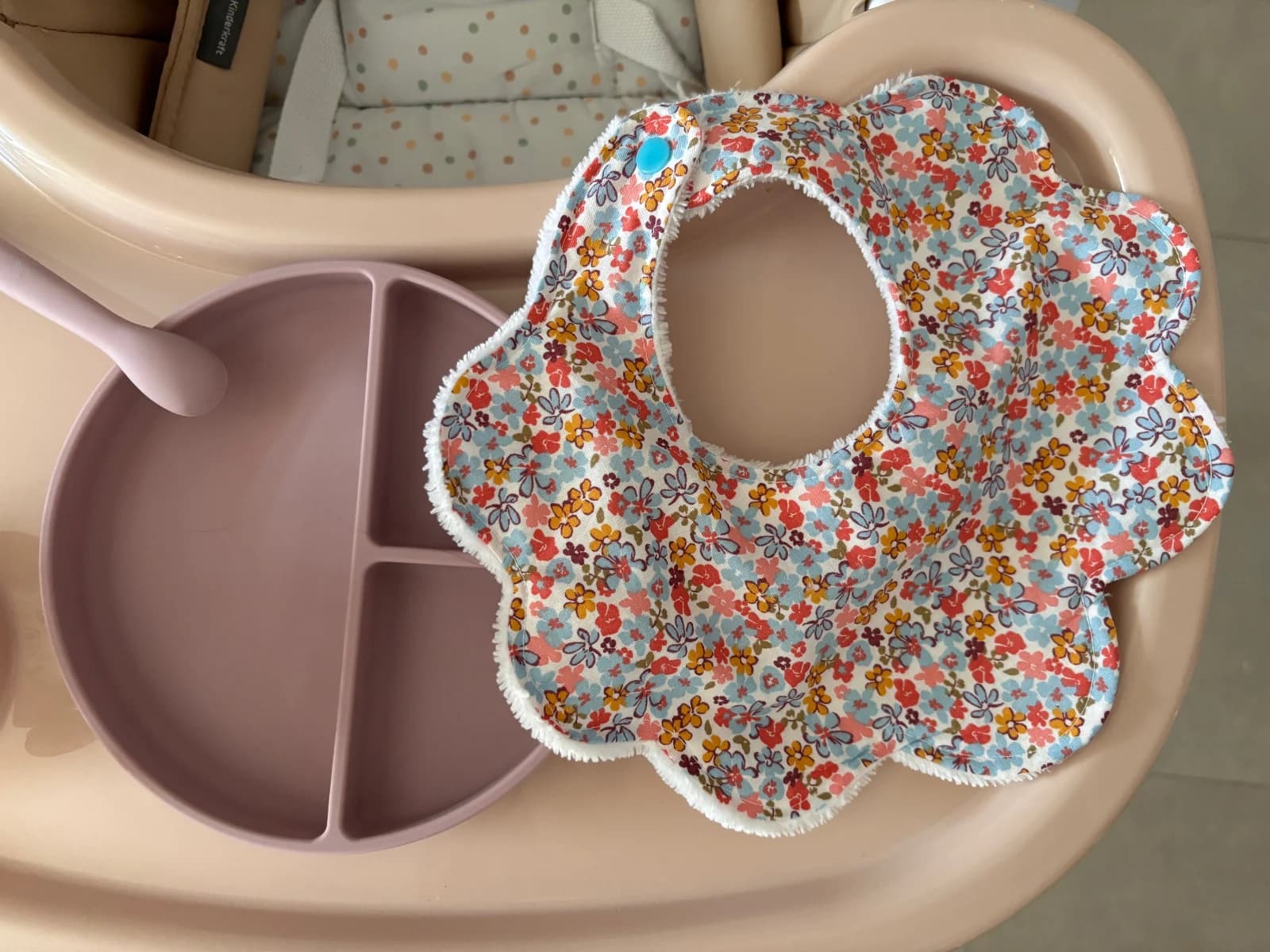 Bavoir pour bébé en forme de fleur aux motifs floraux multicolores posé à côté d'une assiette rose