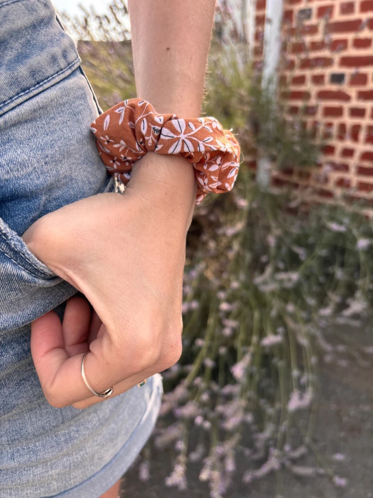 Chouchou en tissu cannelle à motifs floraux blancs porté au poignet devant un mur en briques.