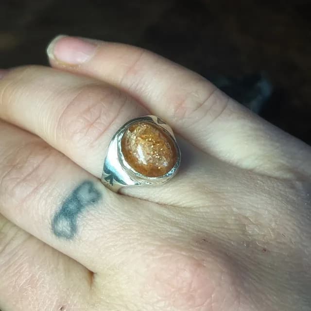 Bague chevalière portée au doigt, sertie d'une pierre cabochon orange scintillante aux reflets dorés.