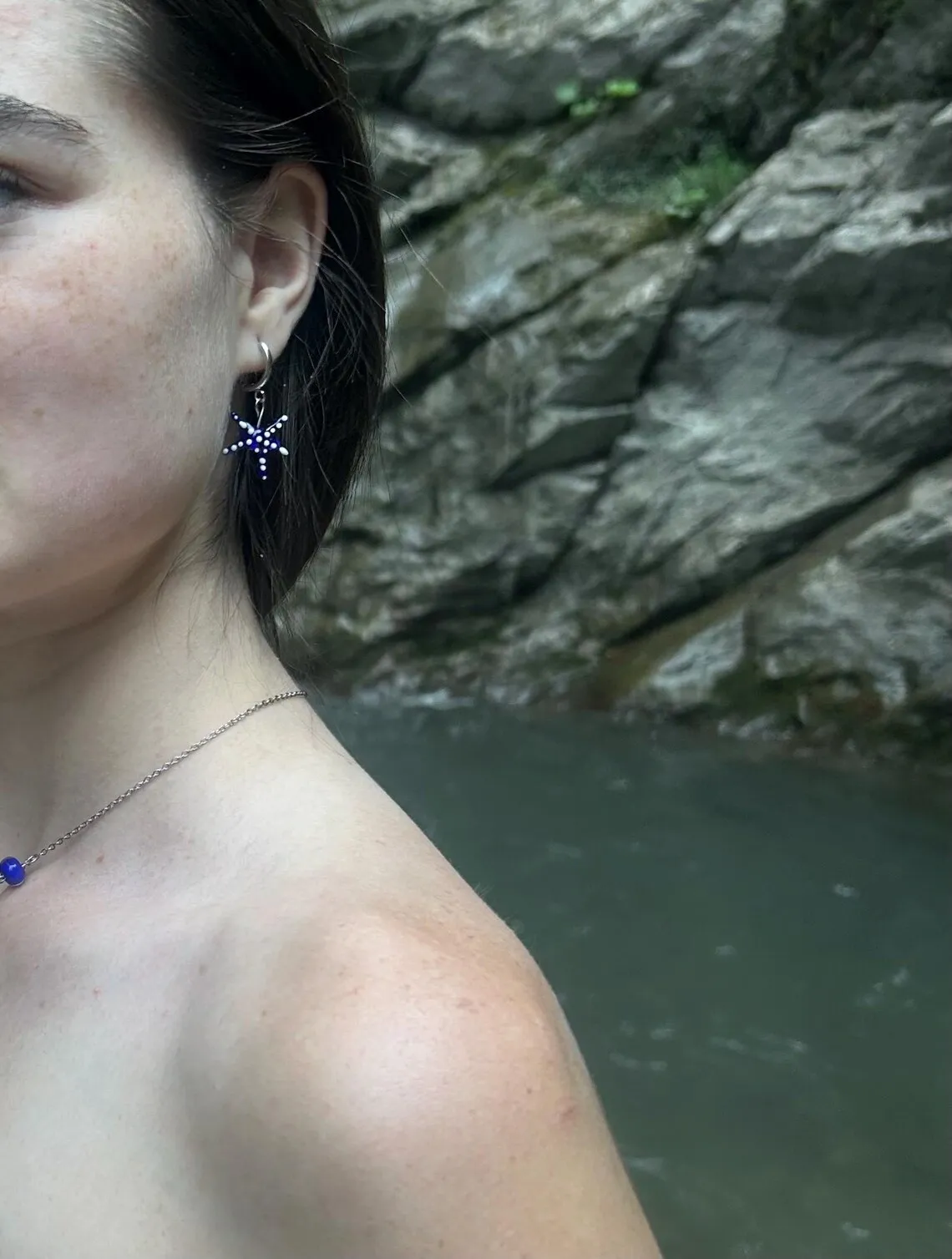Boucle d'oreille portée en forme d'étoile de mer bleue à points blancs sur fond de nature rocheuse.