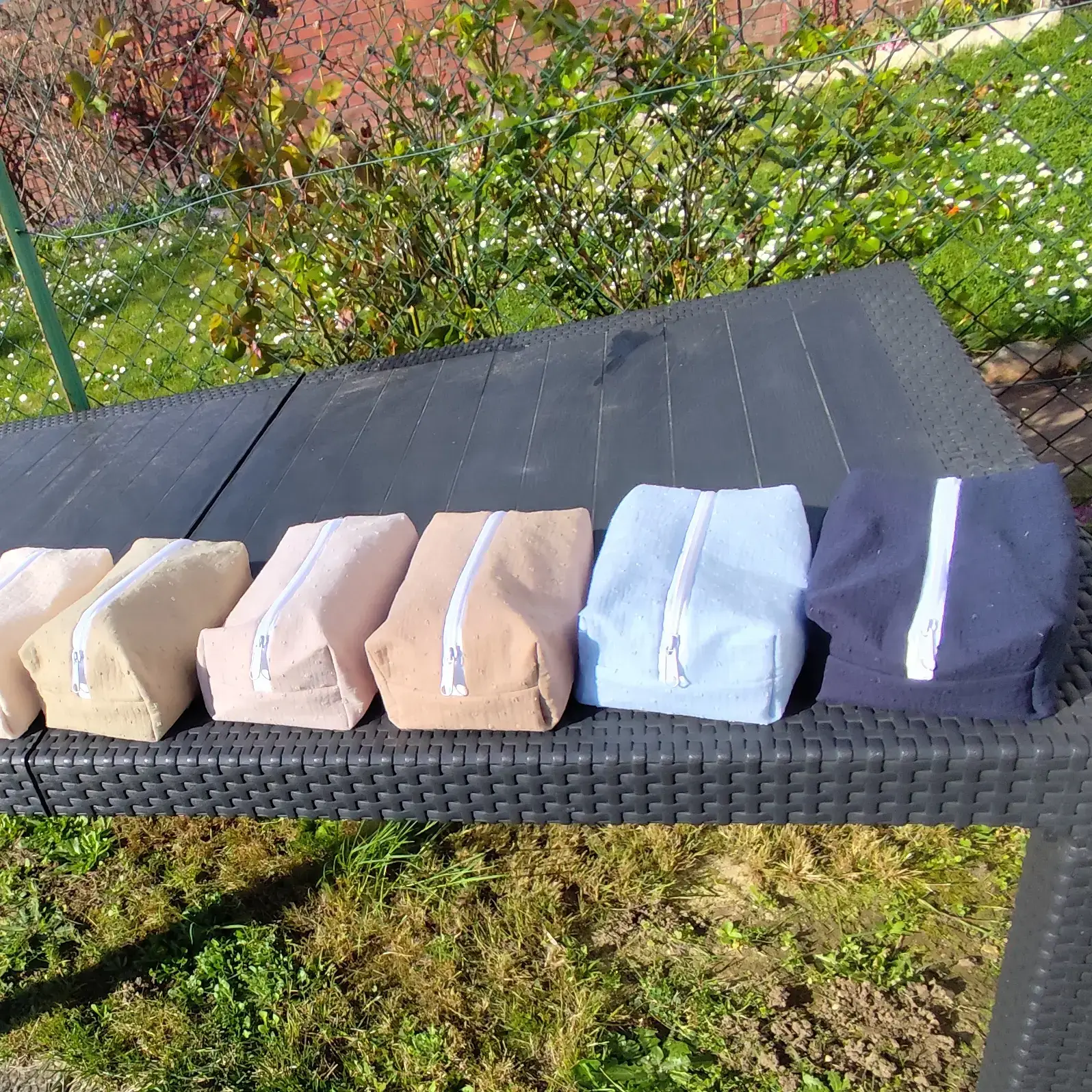 Alignement de six trousses rectangulaires en tissu texturé aux coloris variés posées sur une table d'extérieur