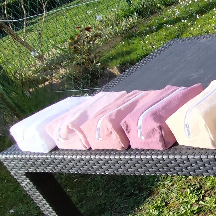 Cinq trousses rectangulaires zippées alignées déclinées en blanc, rose et beige sur une table d'extérieur.
