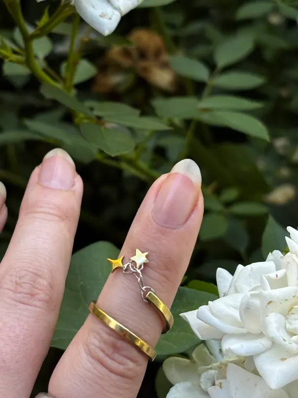 Bague dorée spirale portée au doigt ornée de deux étoiles, devant des roses blanches